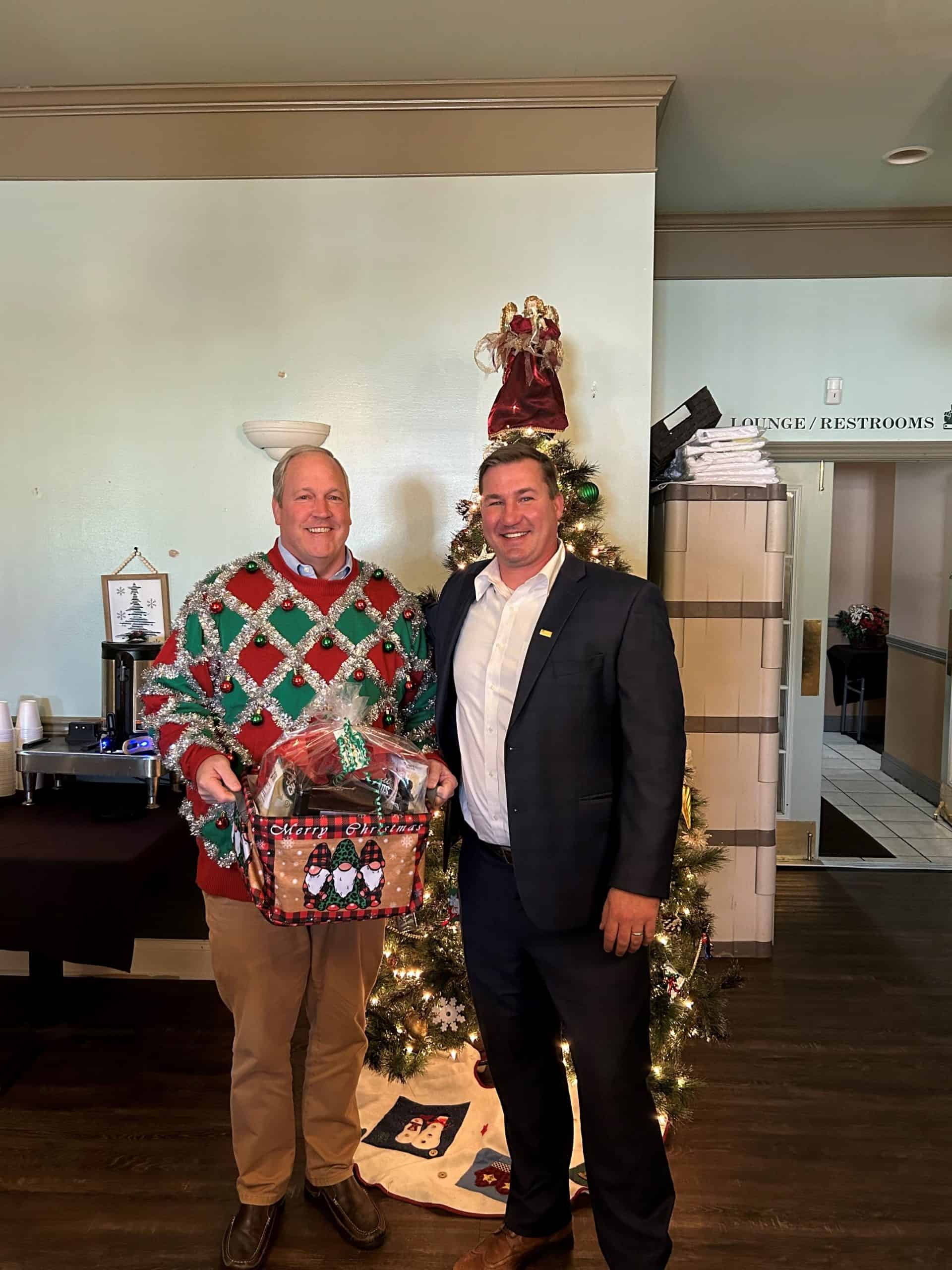 Two men smiling by decorated Christmas tree, one in ugly sweater