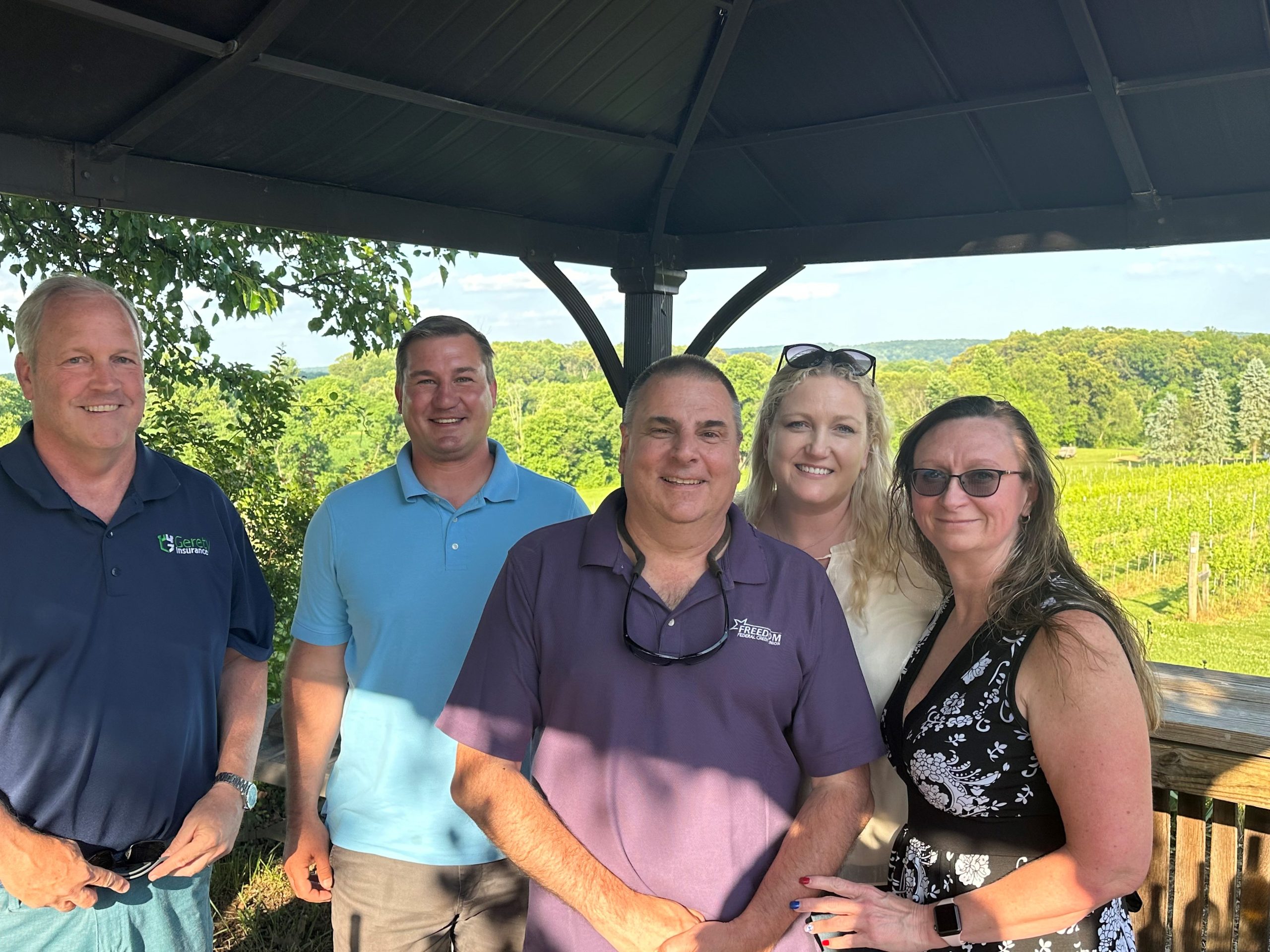 Five adults smiling under a gazebo with green vineyard background