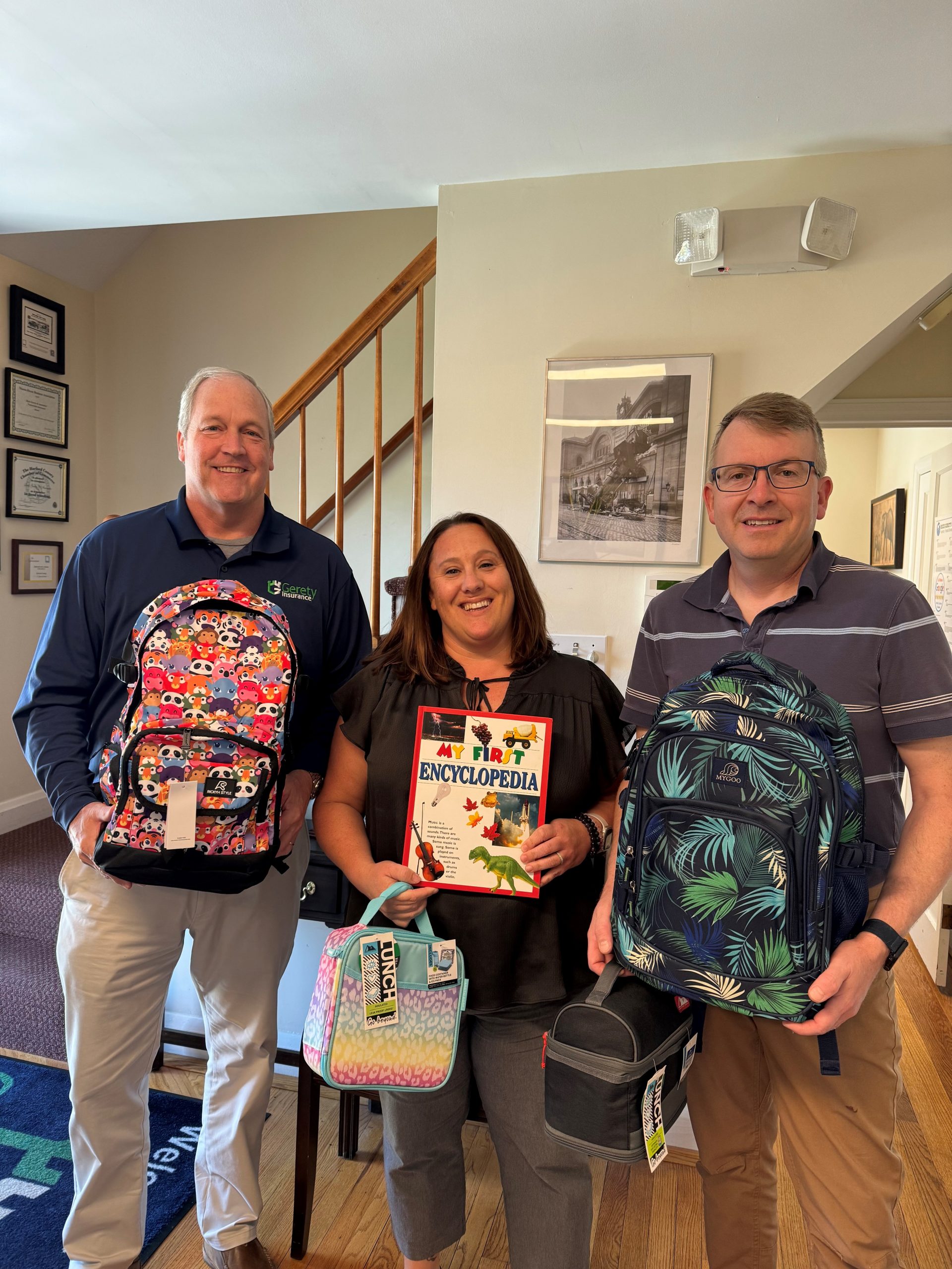 Three Gerety employees holding backpacks, lunch bags, and a children's book
