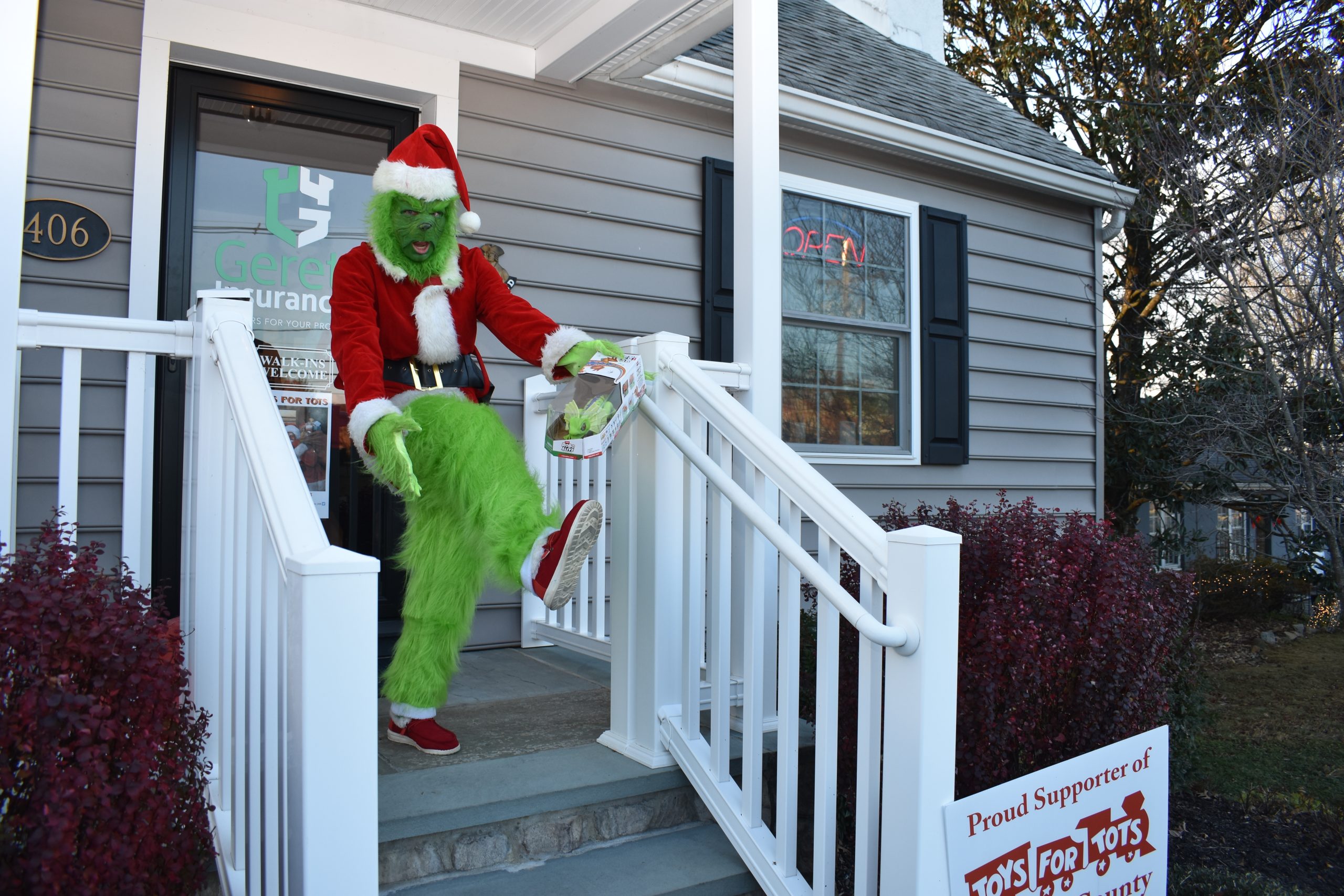 Grinch in Santa suit on porch kicking while holding a toy box
