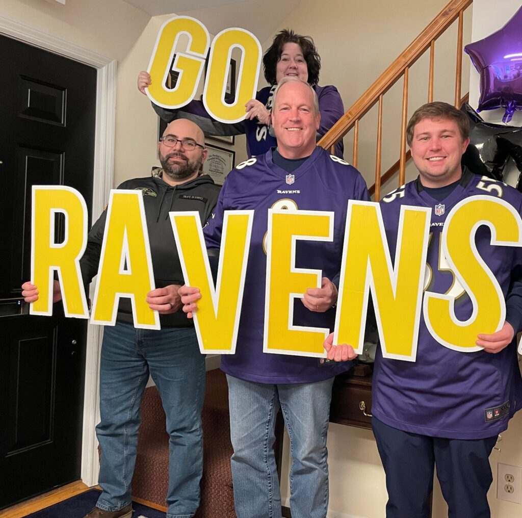 Four fans in purple jerseys holding large yellow letters spelling GO RAVENS.