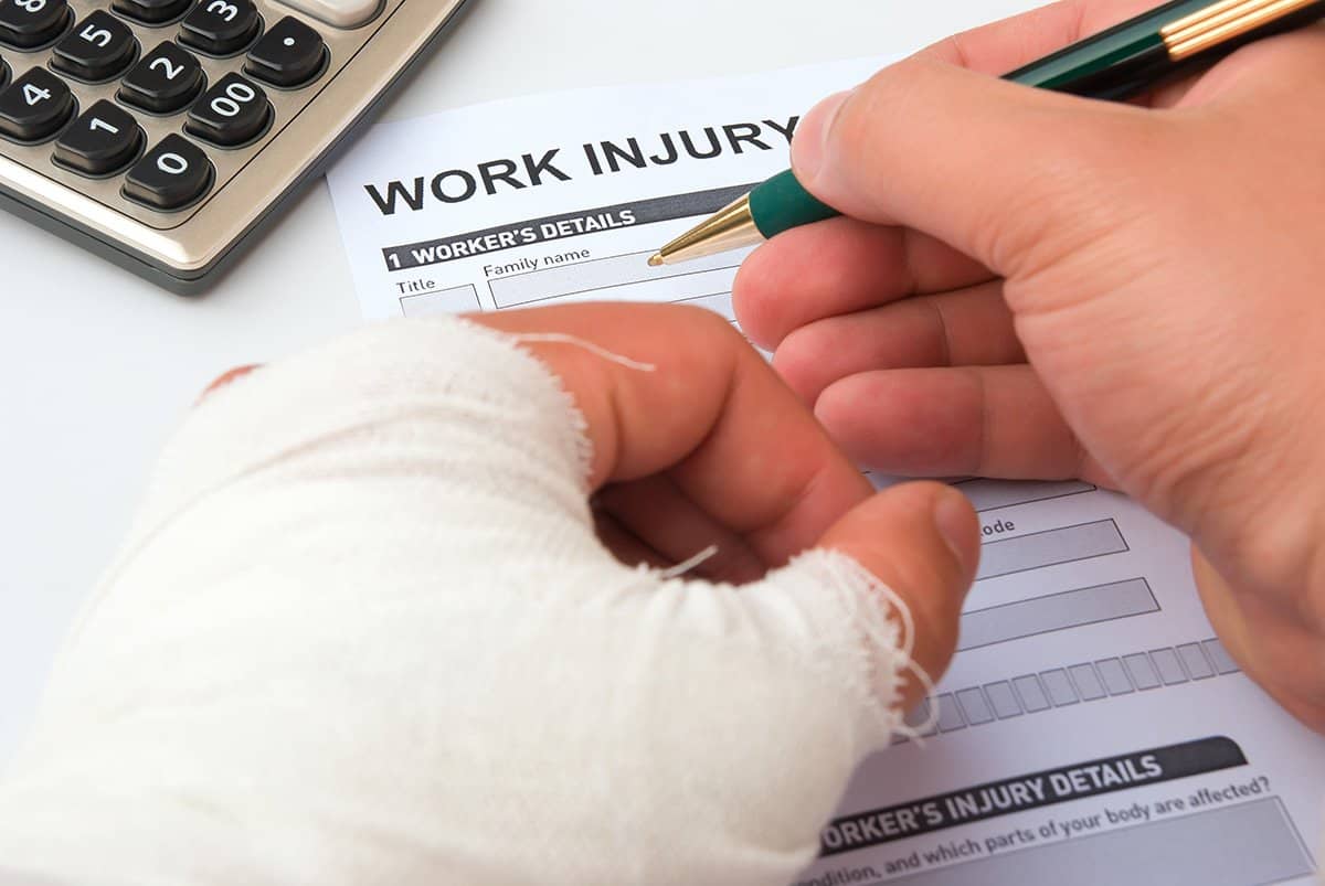 Person with heavily bandaged hand filling out a 'Work Injury' form with pen.