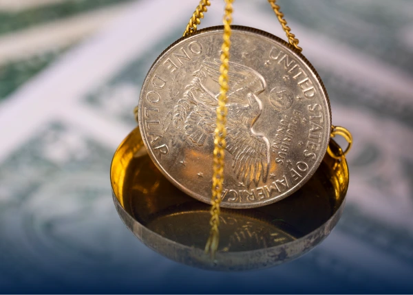 A US silver dollar rests on a small gold scale pan suspended by chains.