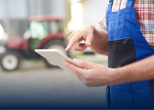 Farm worker in blue overalls tapping a tablet with a tractor blurred behind