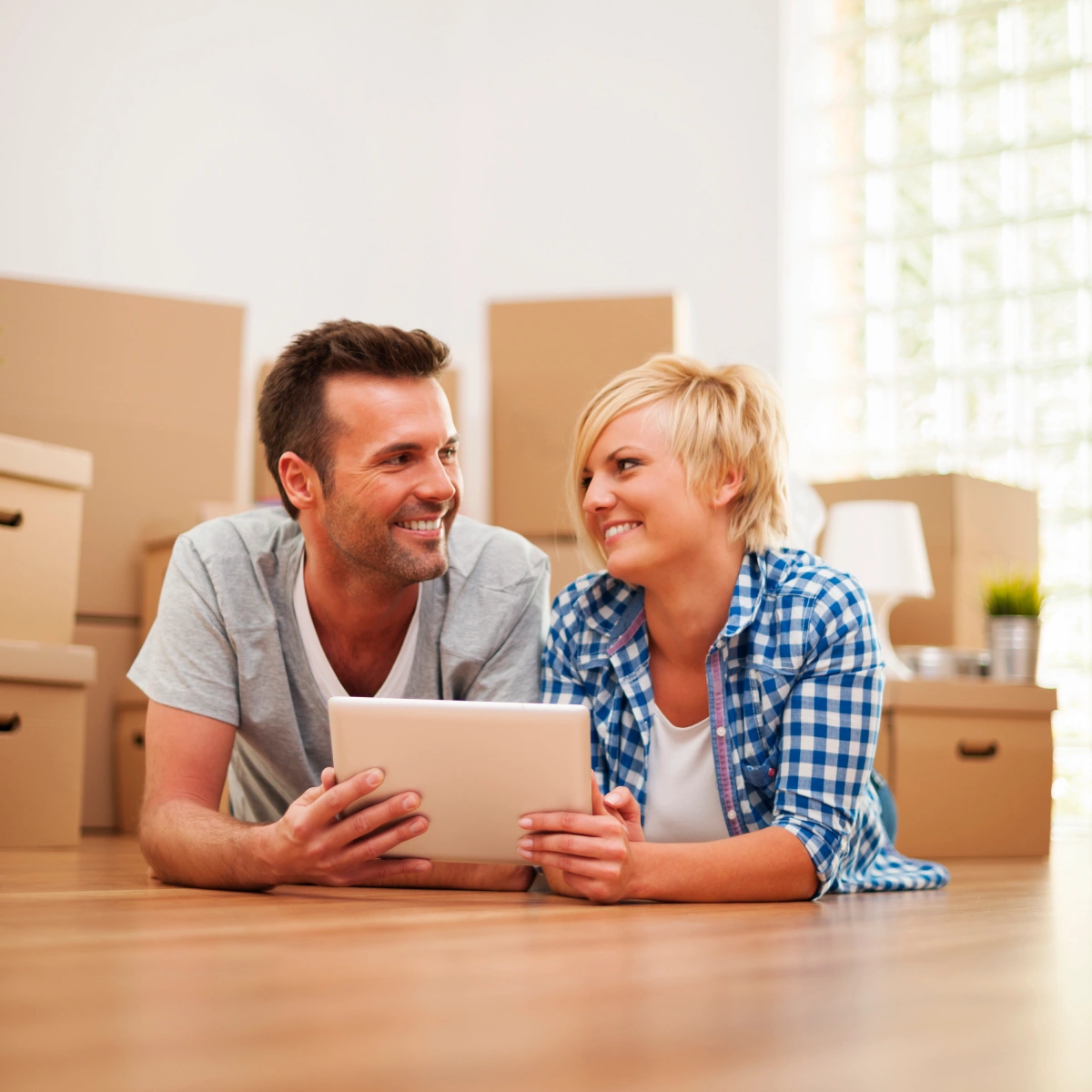 Smiling couple lying on floor using tablet with packed moving boxes in background.