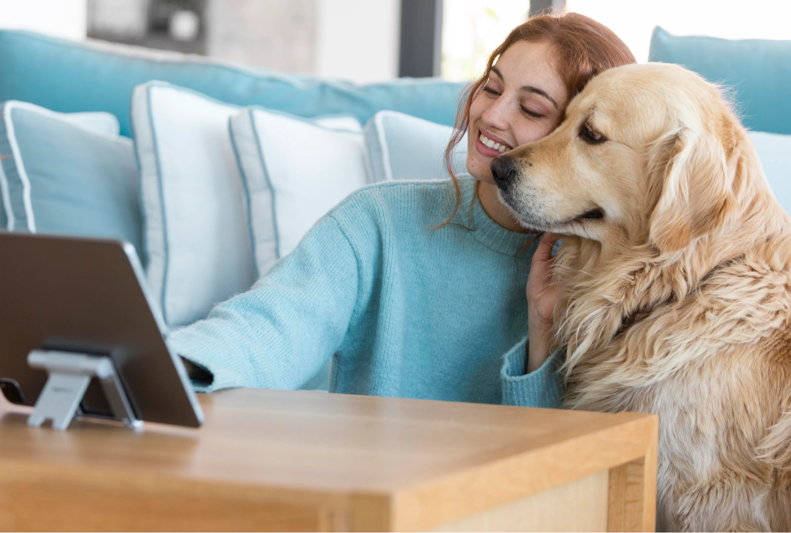 Smiling woman in blue sweater with golden retriever, using tablet on wooden table indoors.