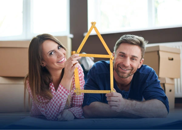 A smiling couple lie on the floor holding a folding ruler shaped like a house.