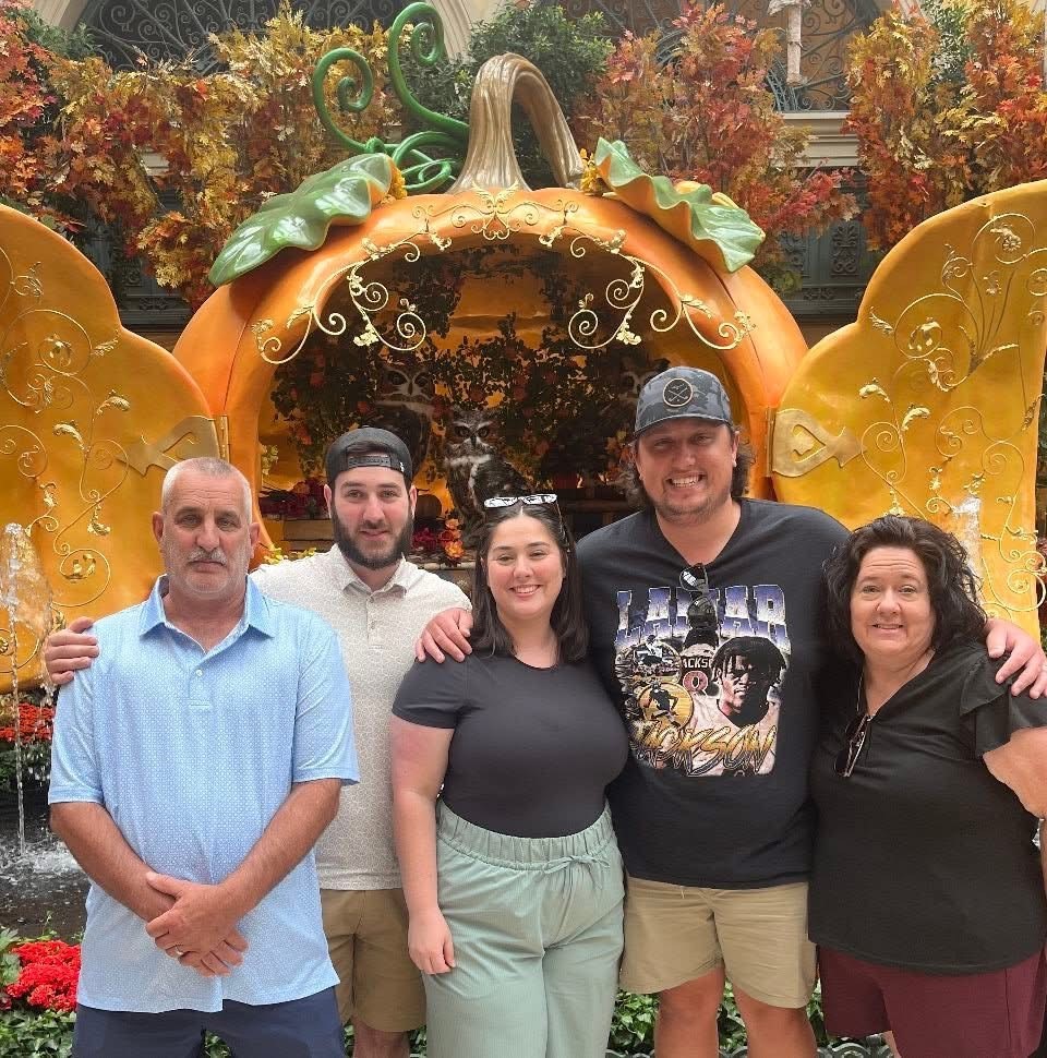 Five people posing in front of a large decorative pumpkin with autumn leaves and flowers.