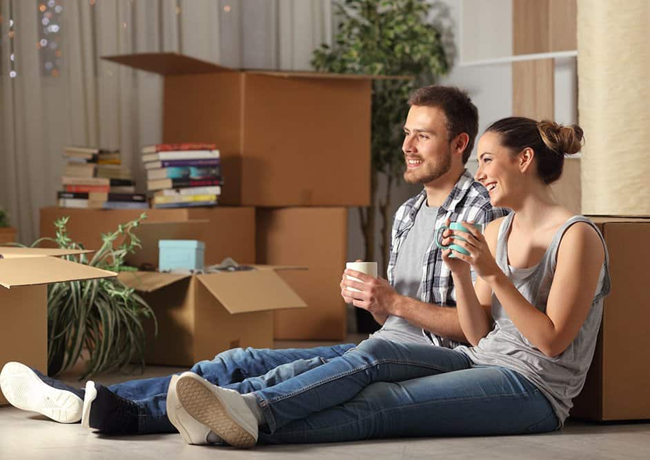 Young couple sitting on floor among moving boxes, smiling and holding mugs