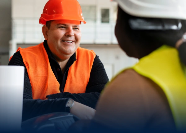 Smiling construction worker in orange hard hat and vest talking to colleague.