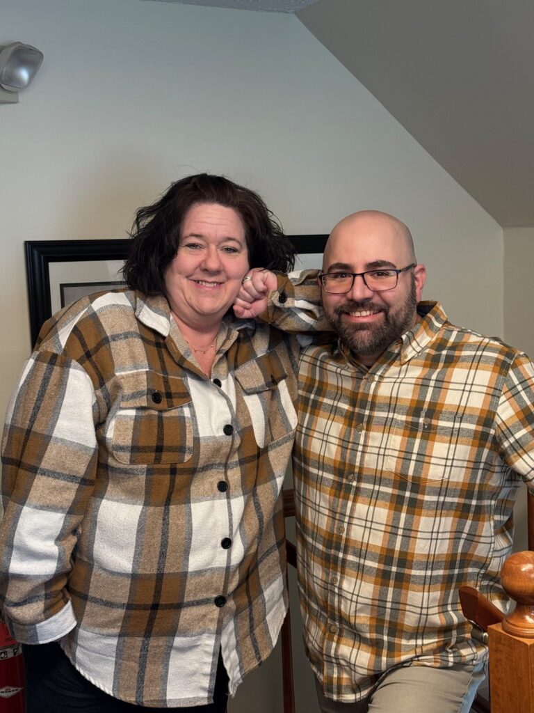 Two smiling adults in brown plaid shirts standing by a stair railing
