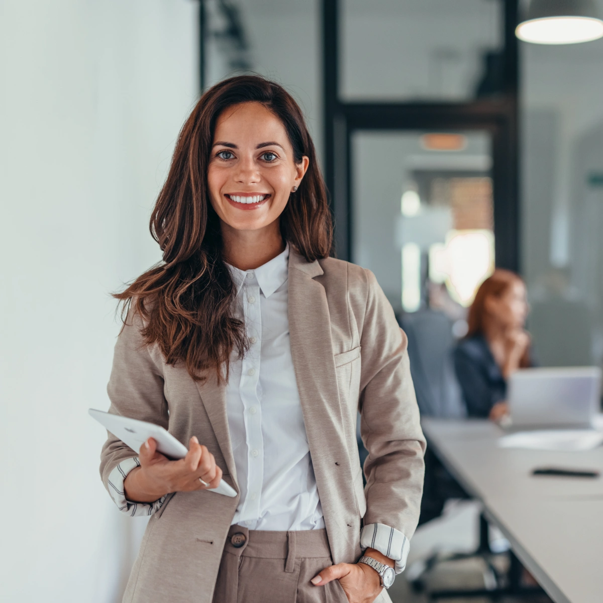 Smiling businesswoman in beige suit holding a tablet in a modern office setting.