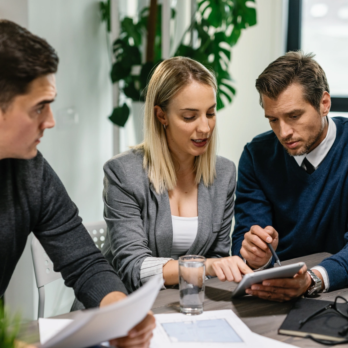 Three coworkers review documents and a tablet together in a modern office setting.