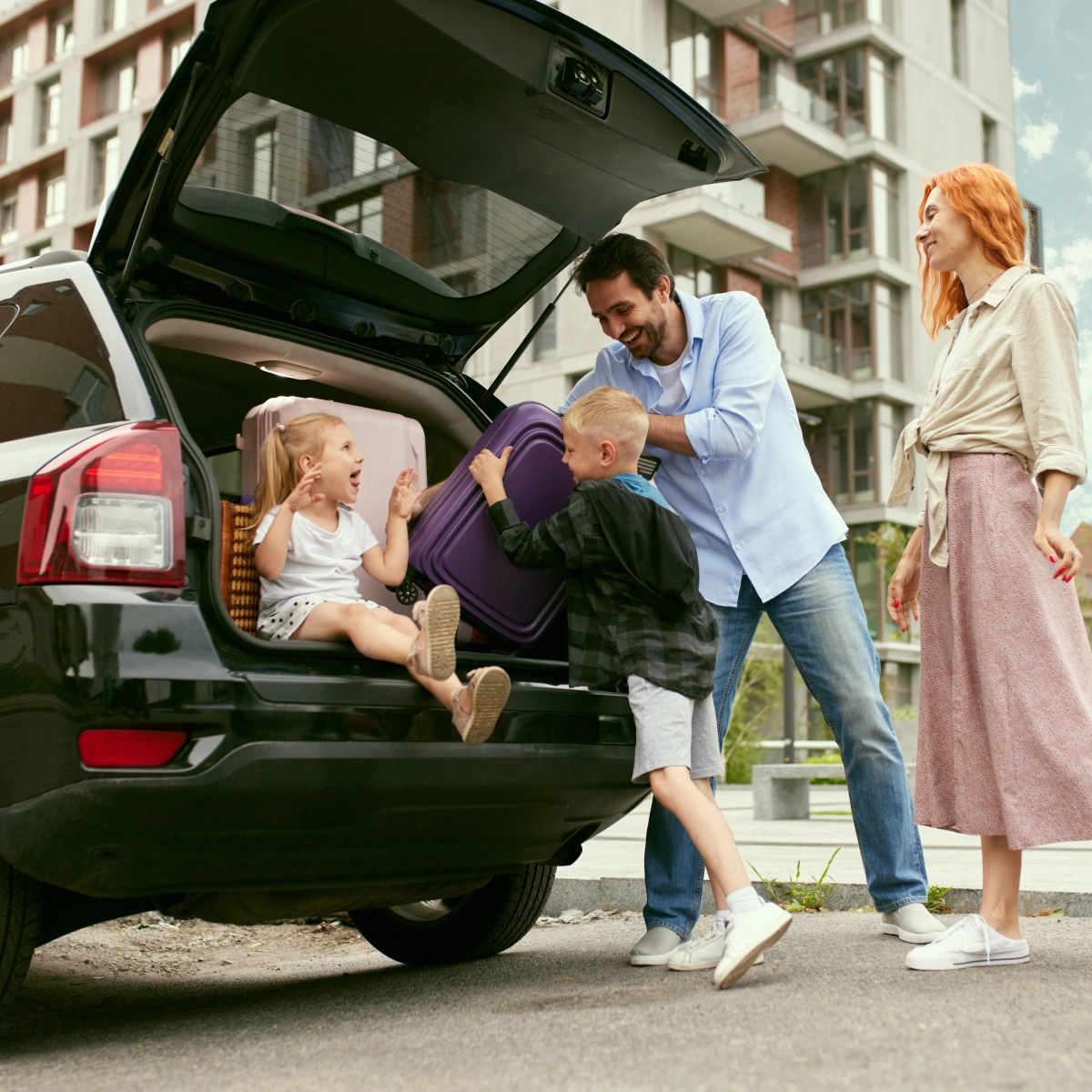 Family packing suitcases into car trunk with daughter sitting inside, preparing for trip outdoors