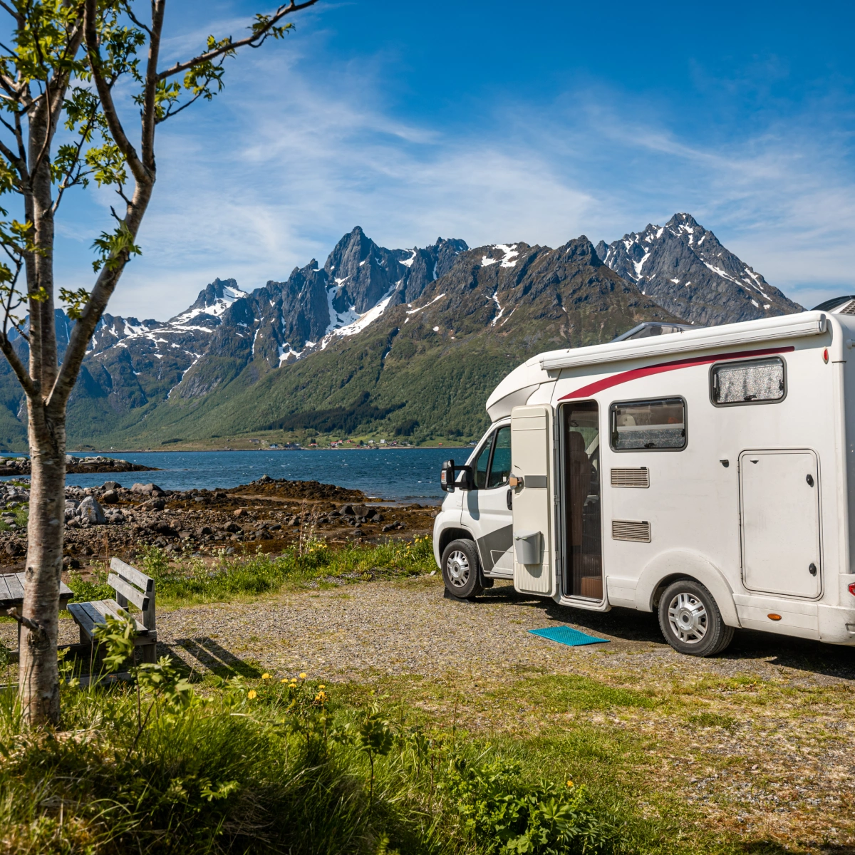 White camper van parked near rocky shore with mountains and blue sky in the background.