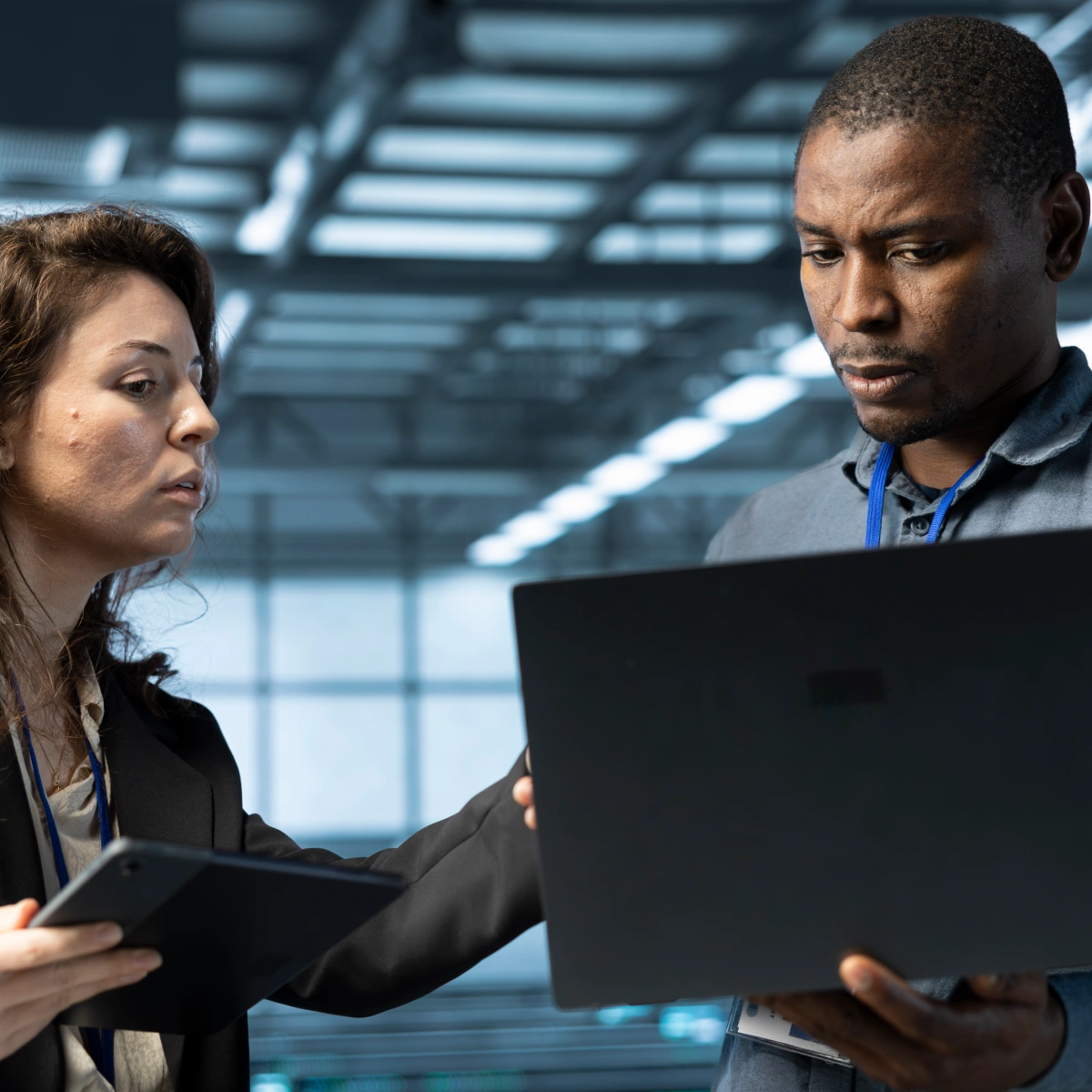 Two coworkers discussing work while one holds a laptop and the other a tablet in an office.