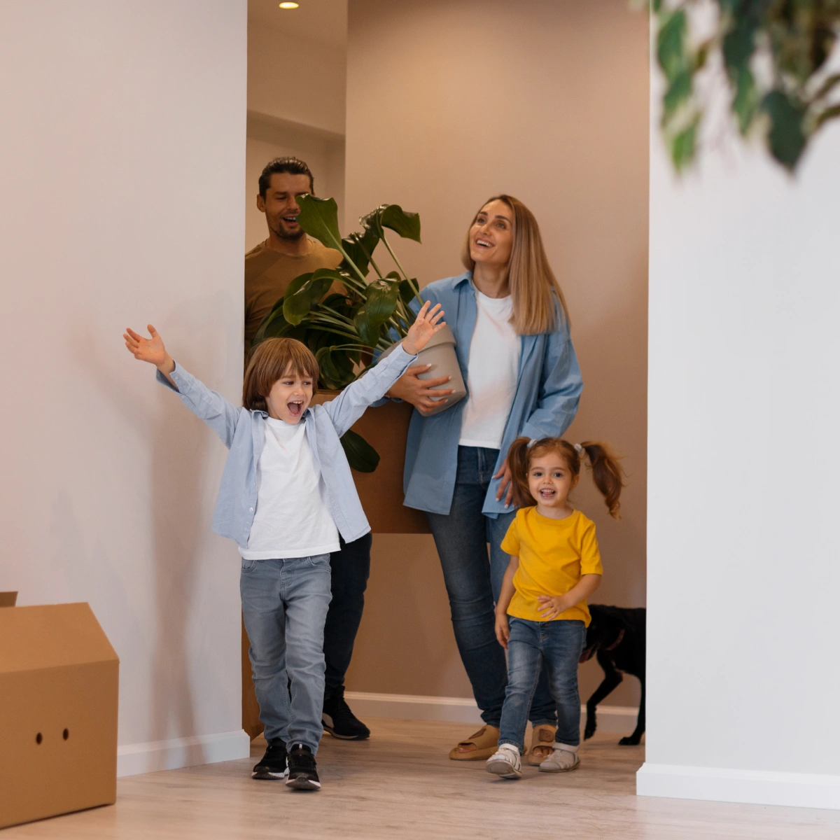 Happy family with two kids and a dog entering a new home carrying a plant and smiling.