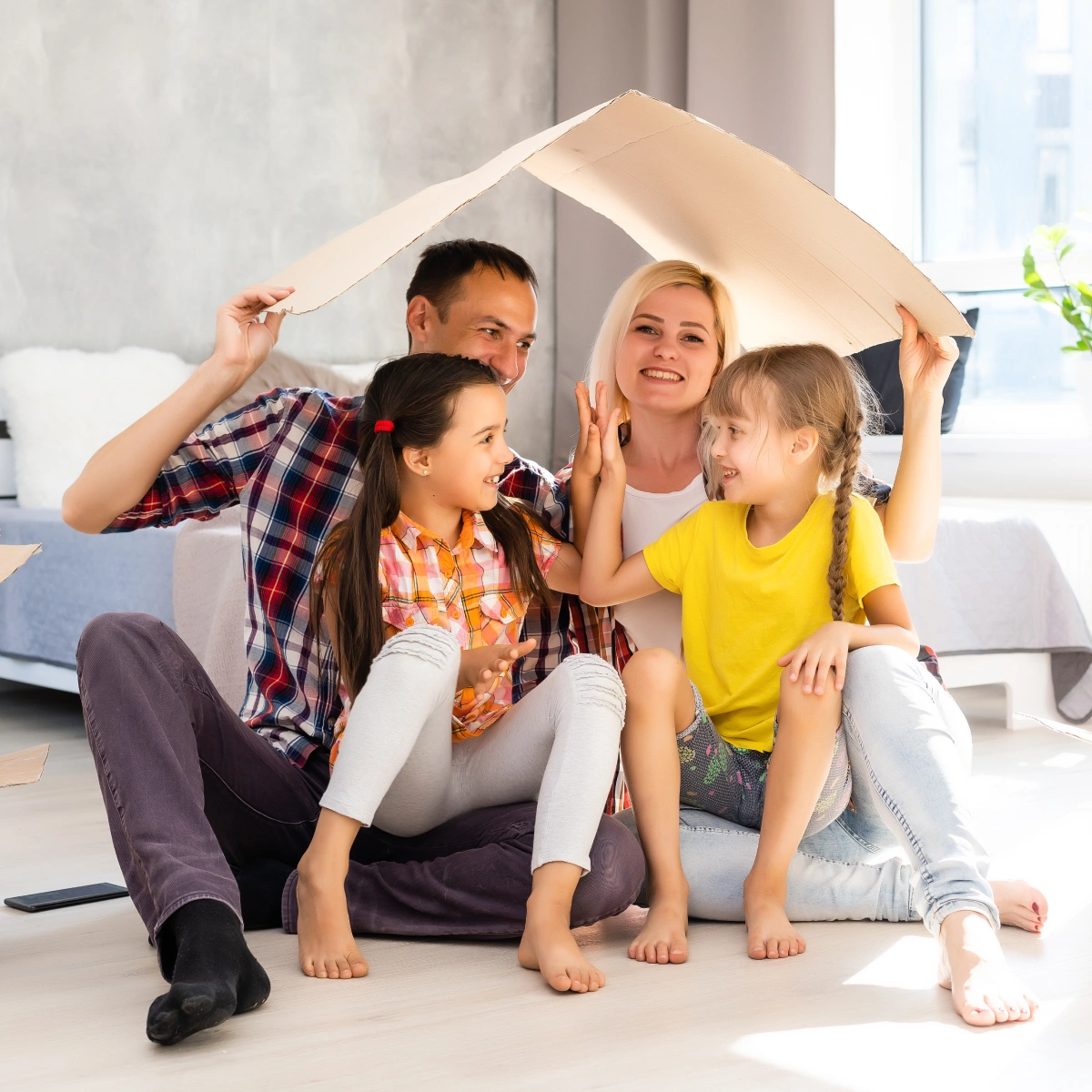 Family of four sitting on floor under cardboard roof, two girls giving high five, smiling