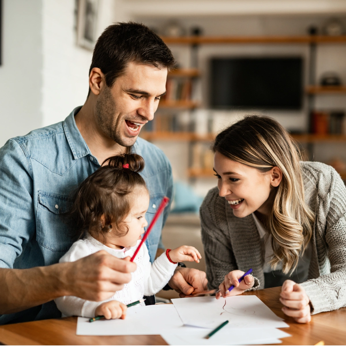 Parents smiling and drawing with their toddler at a table in a cozy living room.