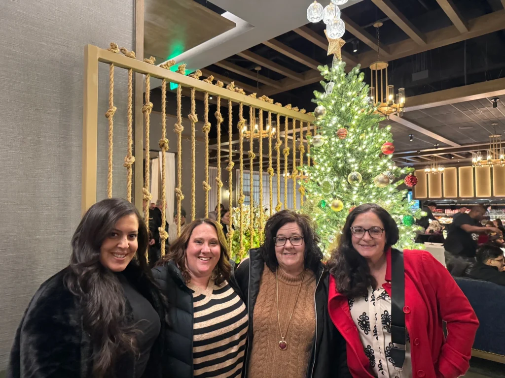 Four women smiling in front of a decorated Christmas tree indoors with warm lighting.